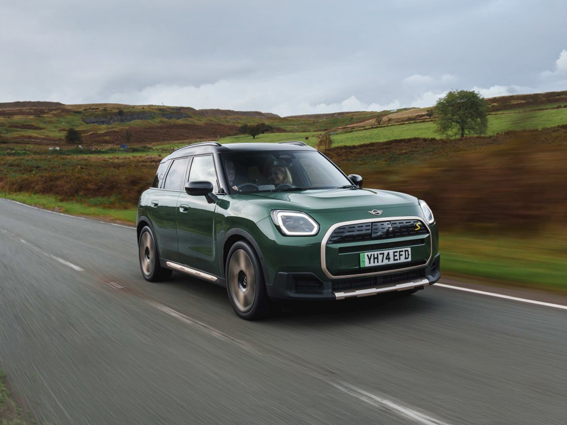 Side view of a driving all-electric MINI Countryman in Smokey Green with Vibrant Silver Roof off-road.
